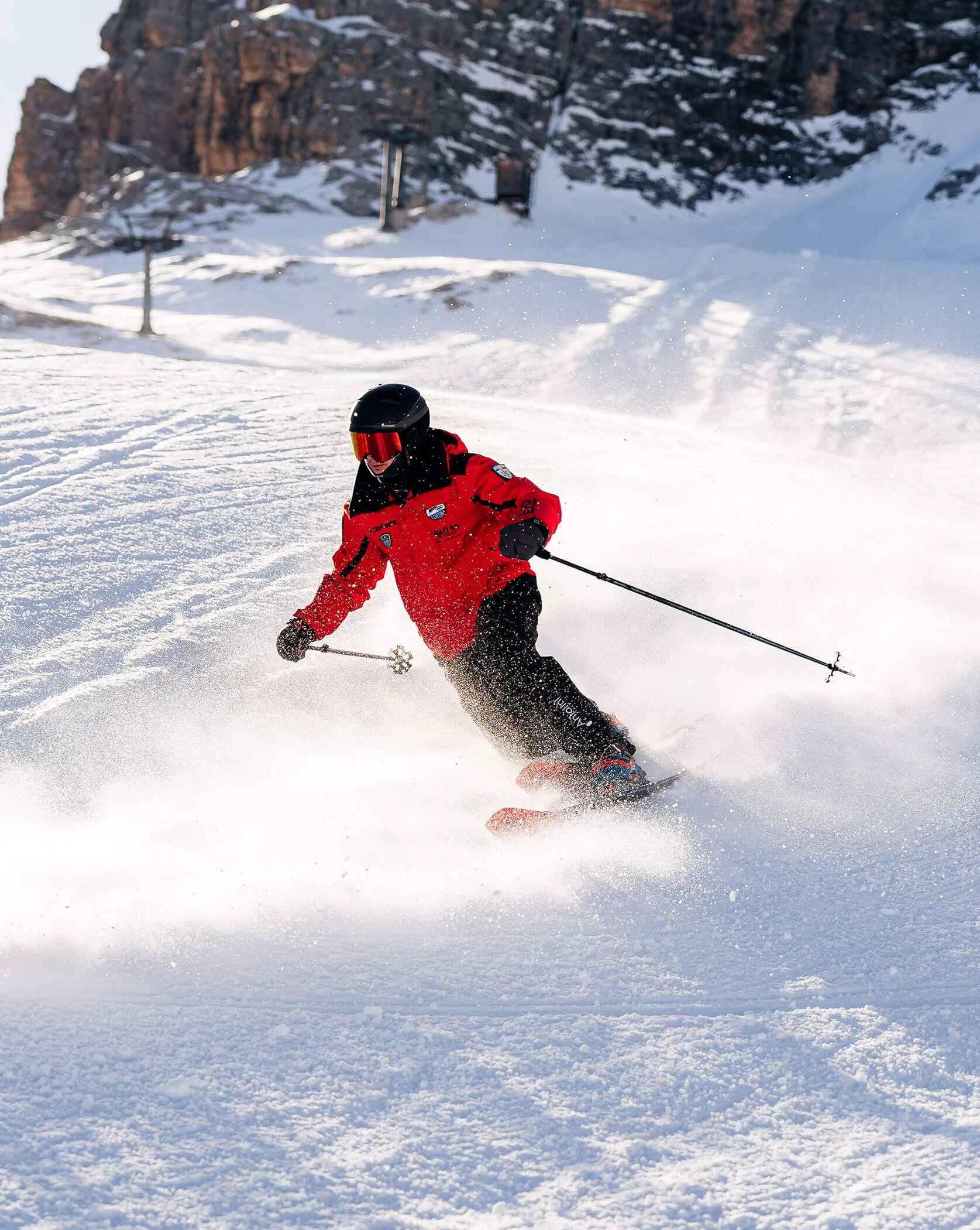 A skier in a red jacket and black helmet skis downhill, kicking up snow on a sunny mountain slope. - Scuola Sci Cortina