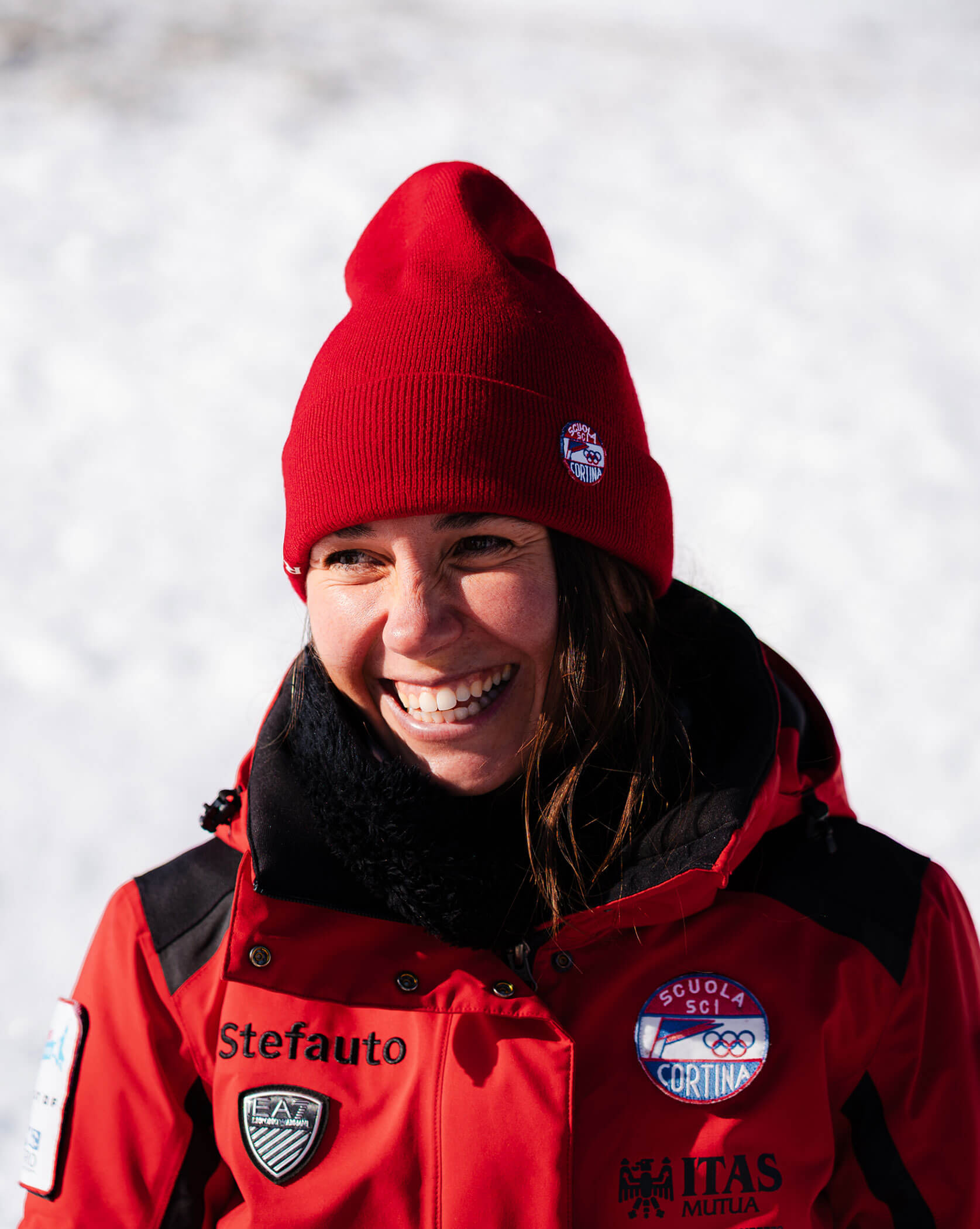 Smiling person in a red jacket and beanie stands outside in a snowy environment, with badges on the jacket. - Scuola Sci Cortina