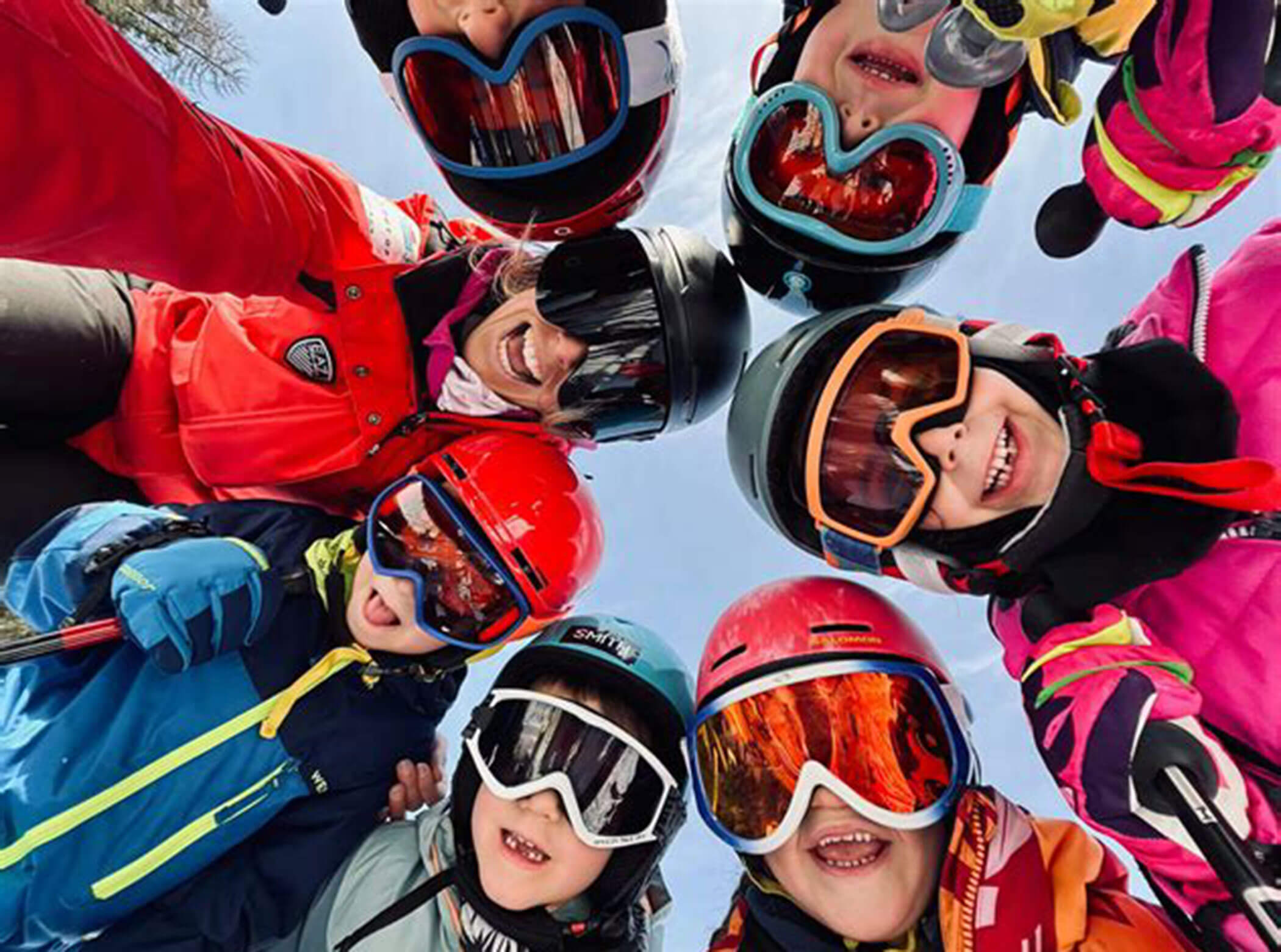 Group of smiling kids and an adult in ski gear, huddled together and looking up at the camera outdoors. - Scuola Sci Cortina