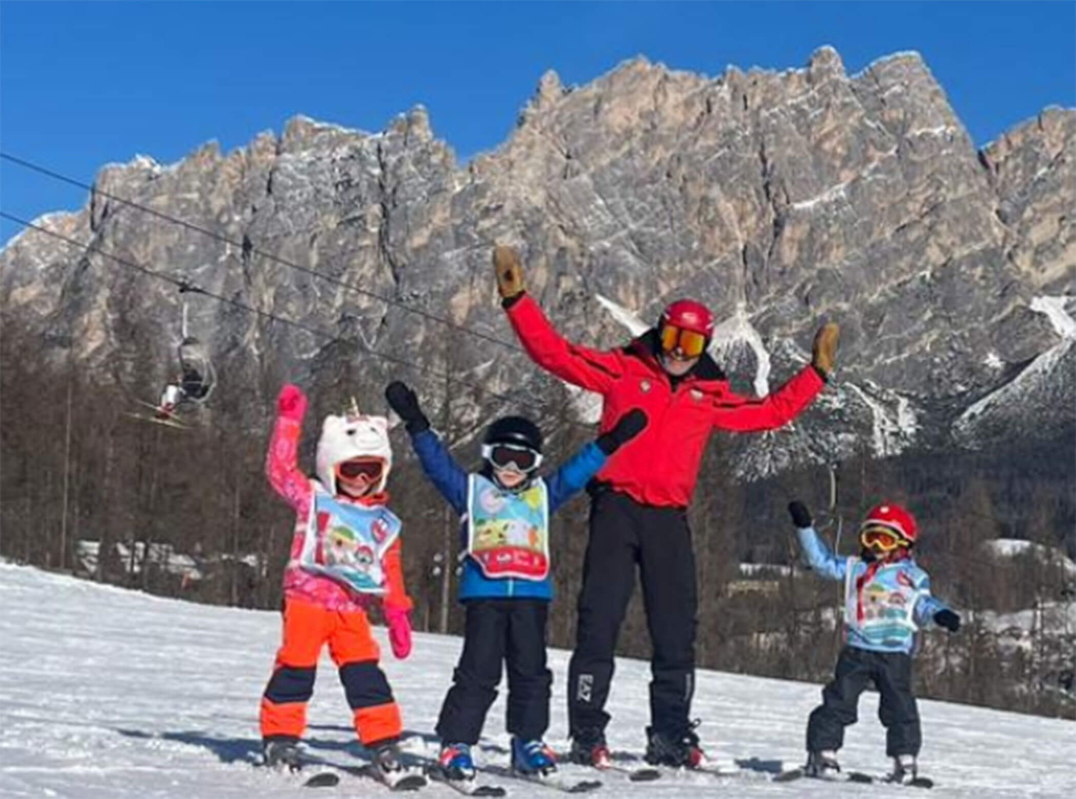 An adult and three children in ski gear pose with raised arms on a snowy mountain slope. - Scuola Sci Cortina