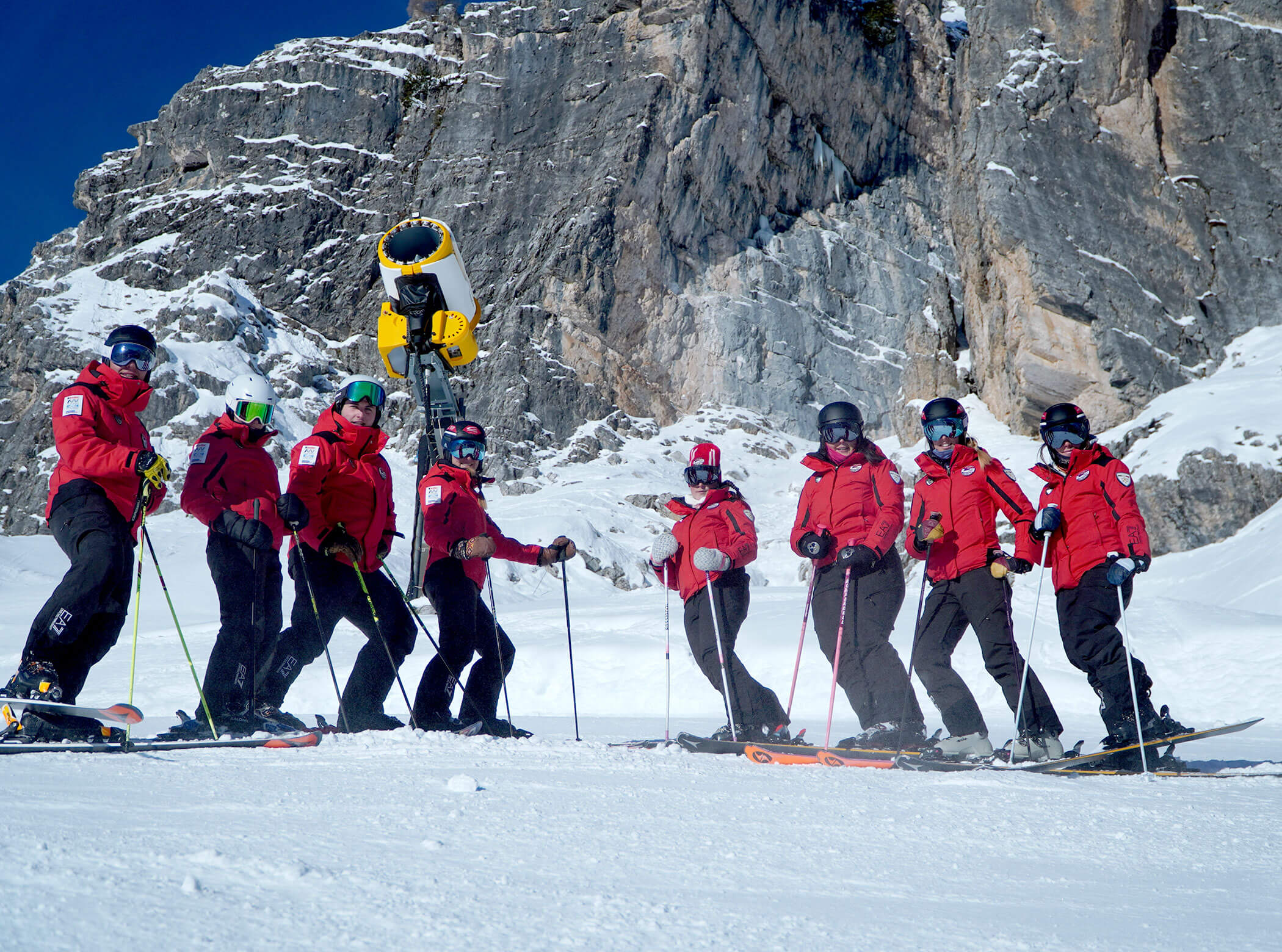 Eight people in red jackets stand on snow with skis, in front of a rocky mountain backdrop. - Scuola Sci Cortina