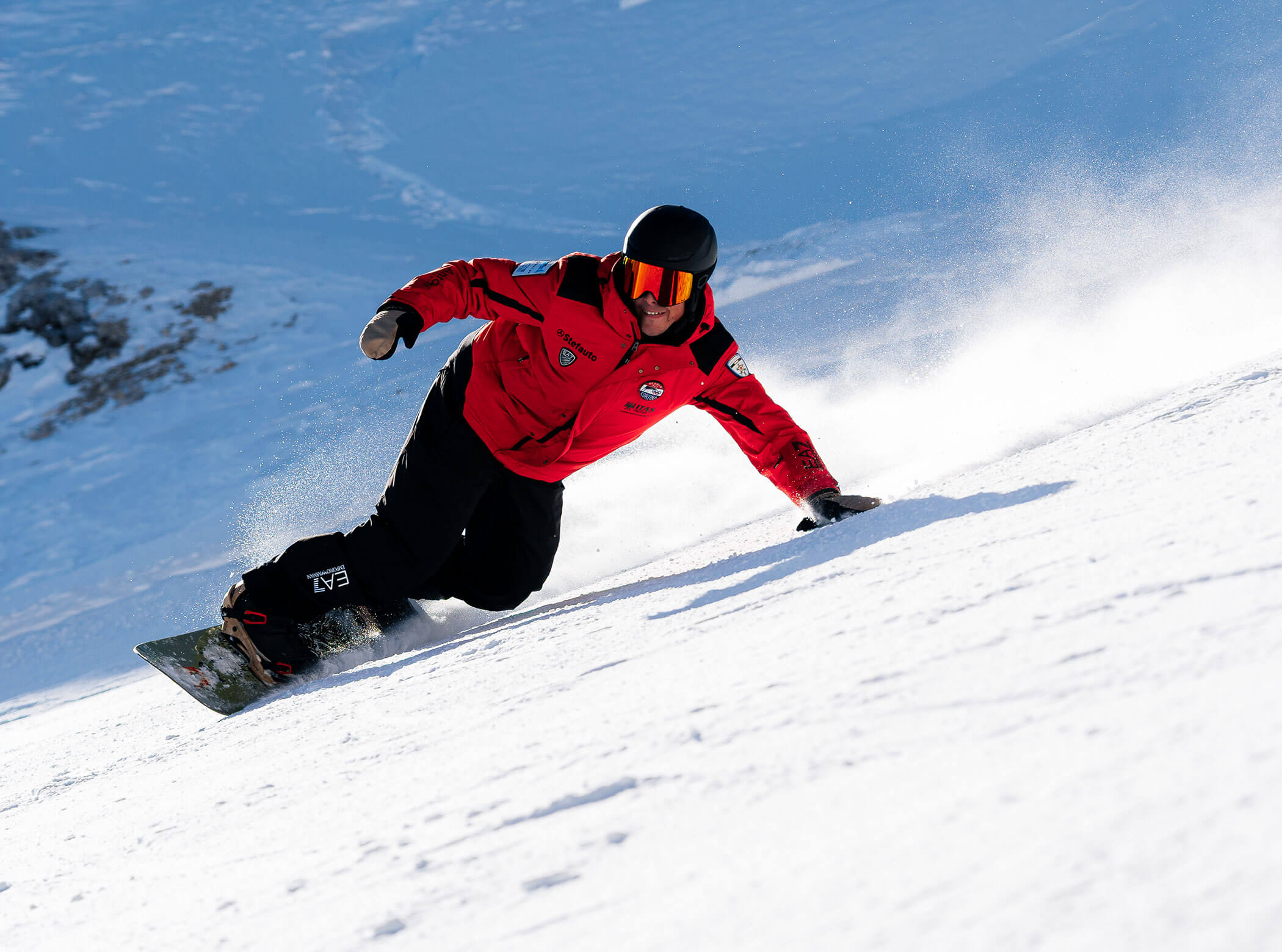 Snowboarder in a red jacket carving down a snowy slope, kicking up powder under a clear sky. - Scuola Sci Cortina