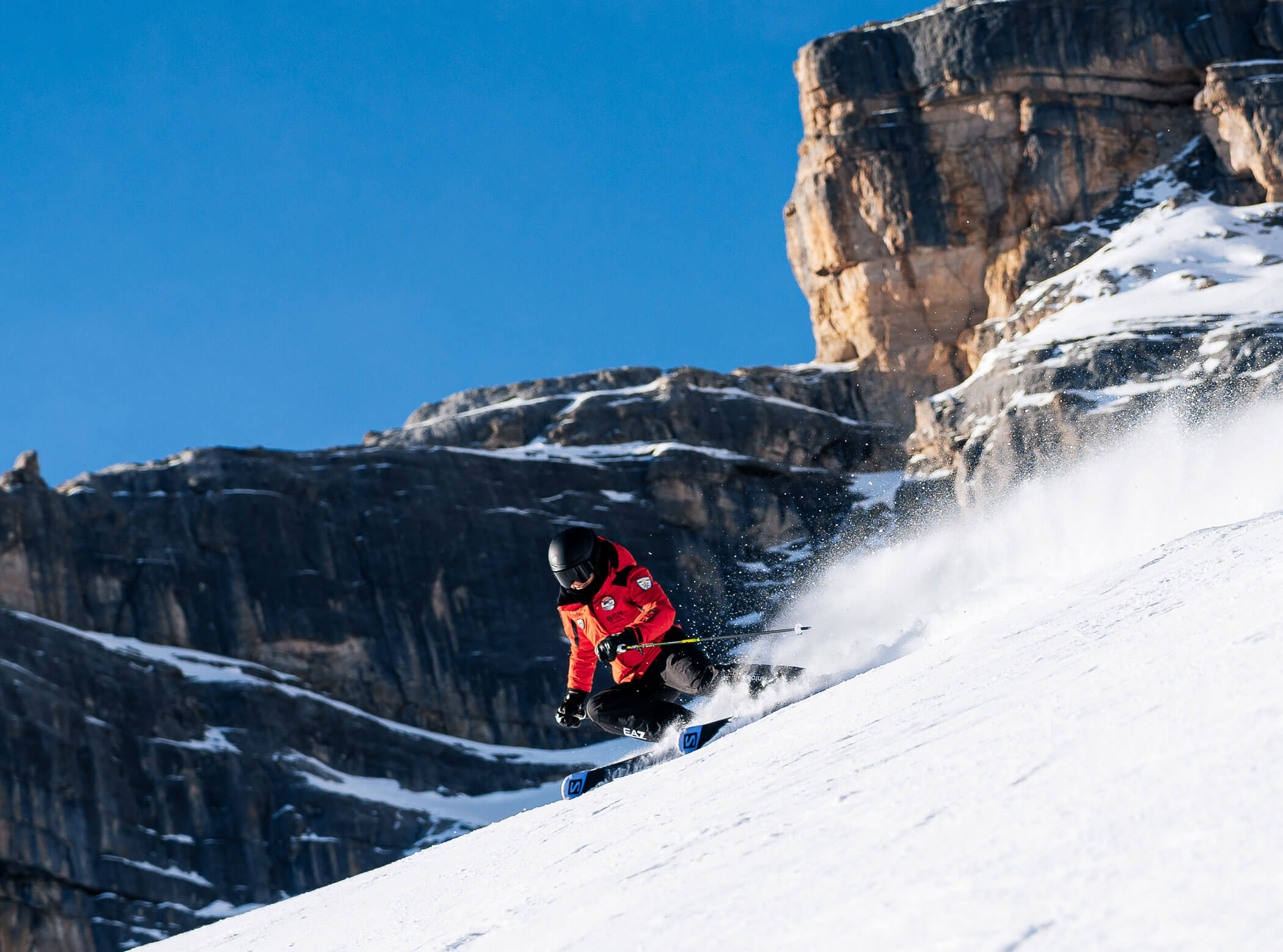 Person skiing downhill on snowy slope with rocky cliffs and blue sky in the background. - Scuola Sci Cortina