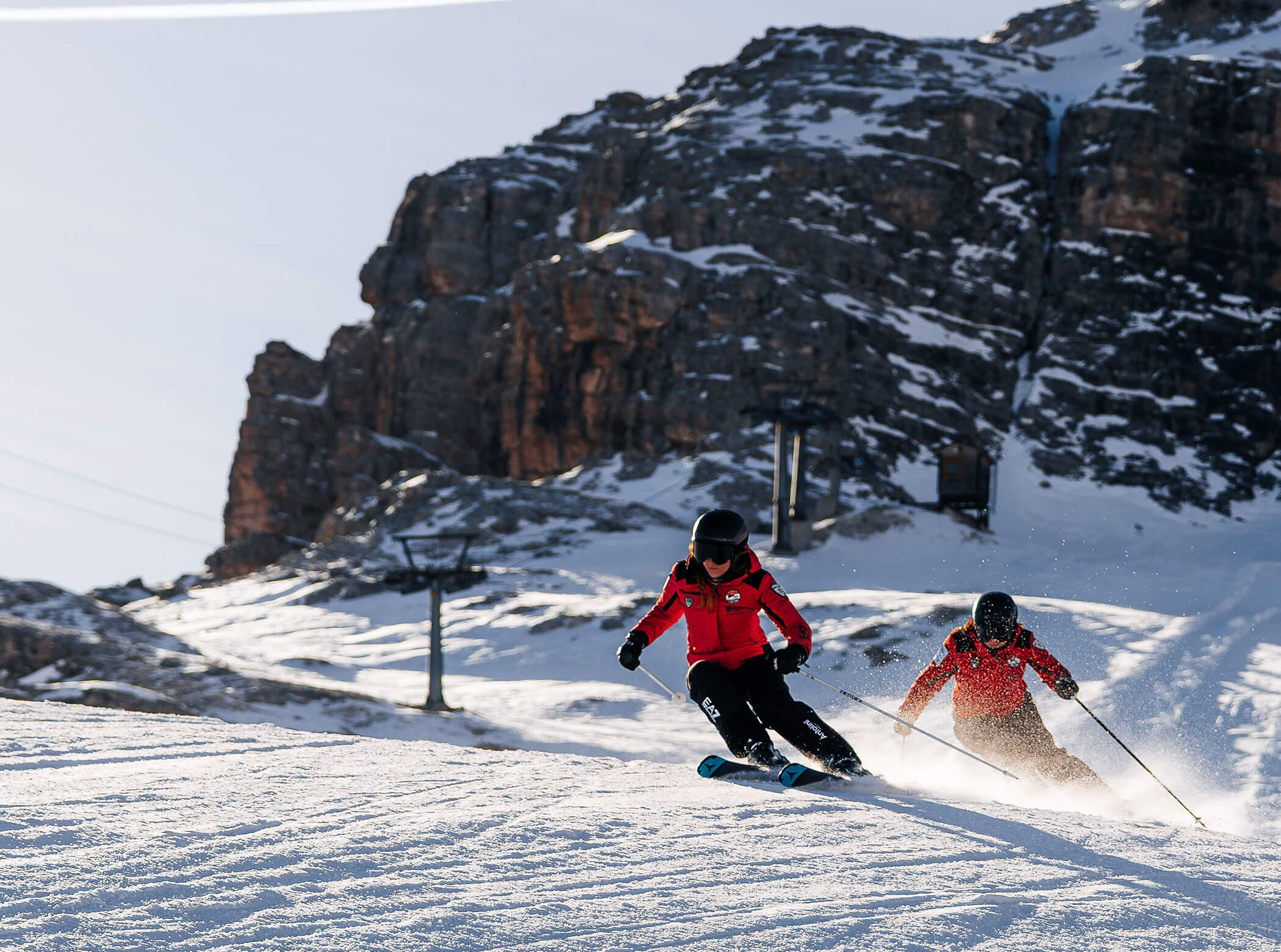 Two skiers in red jackets skiing downhill on a snowy slope with rocky mountains in the background. - Scuola Sci Cortina