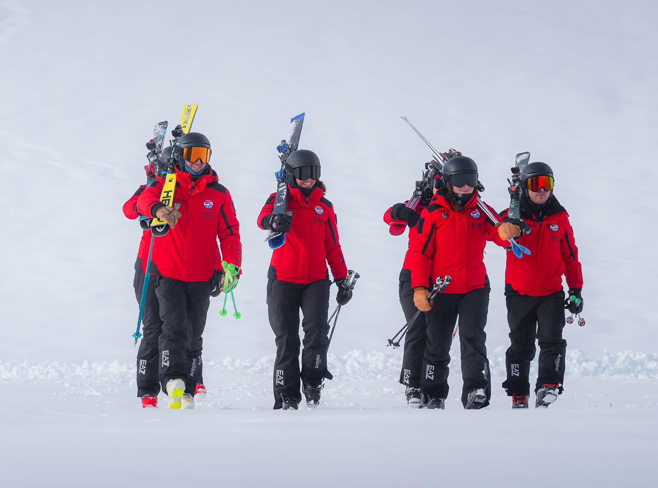 Five people in red jackets and black helmets walk through snow carrying skis and poles. - Scuola Sci Cortina