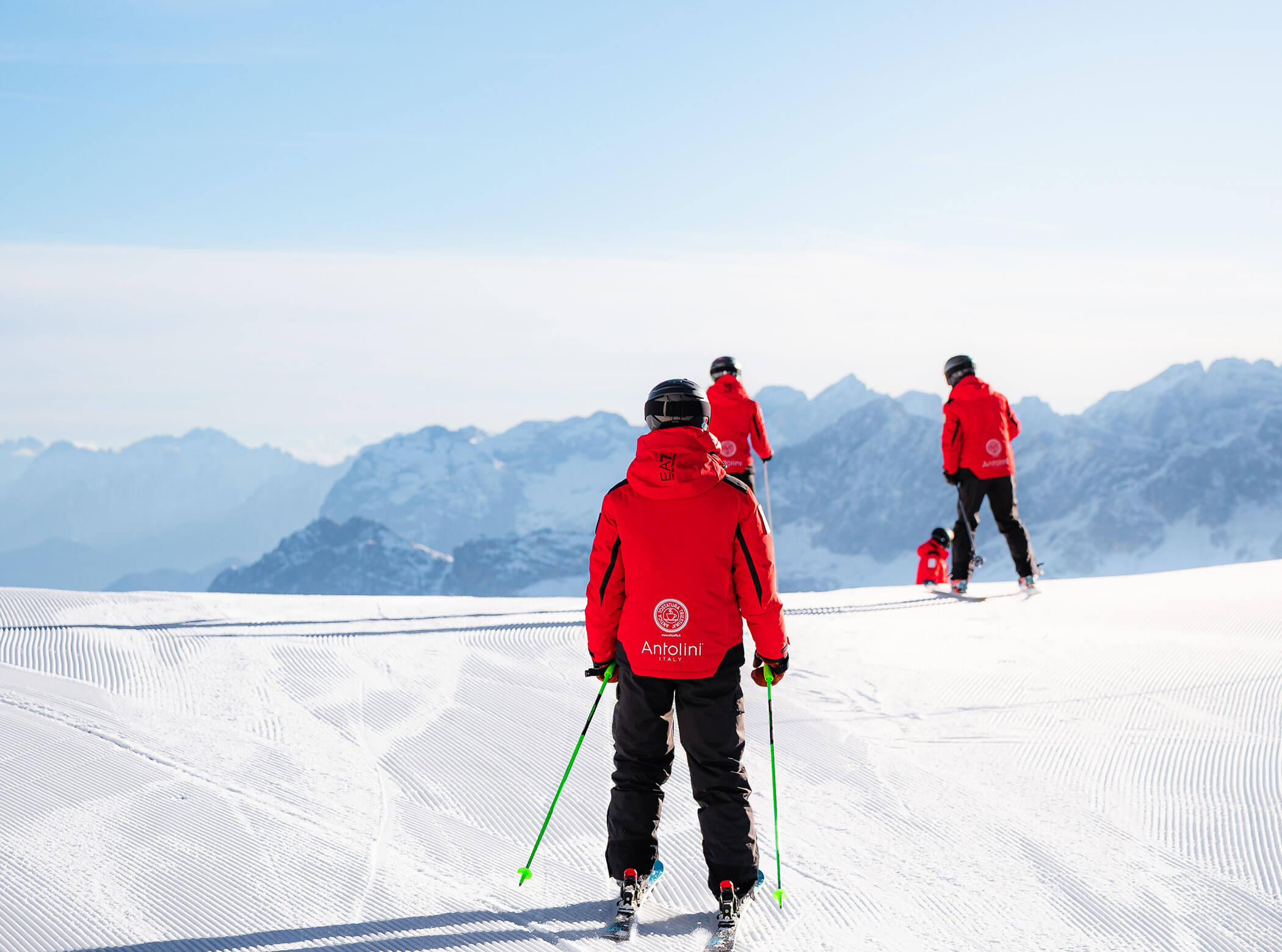 Four skiers in red jackets ski down a snowy slope with mountains visible in the background. - Scuola Sci Cortina