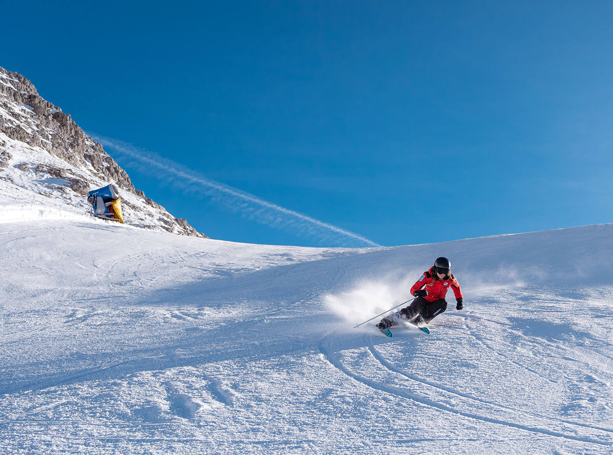 A skier in a red jacket carves down a snowy slope under a clear blue sky with mountains in the background. - Scuola Sci Cortina