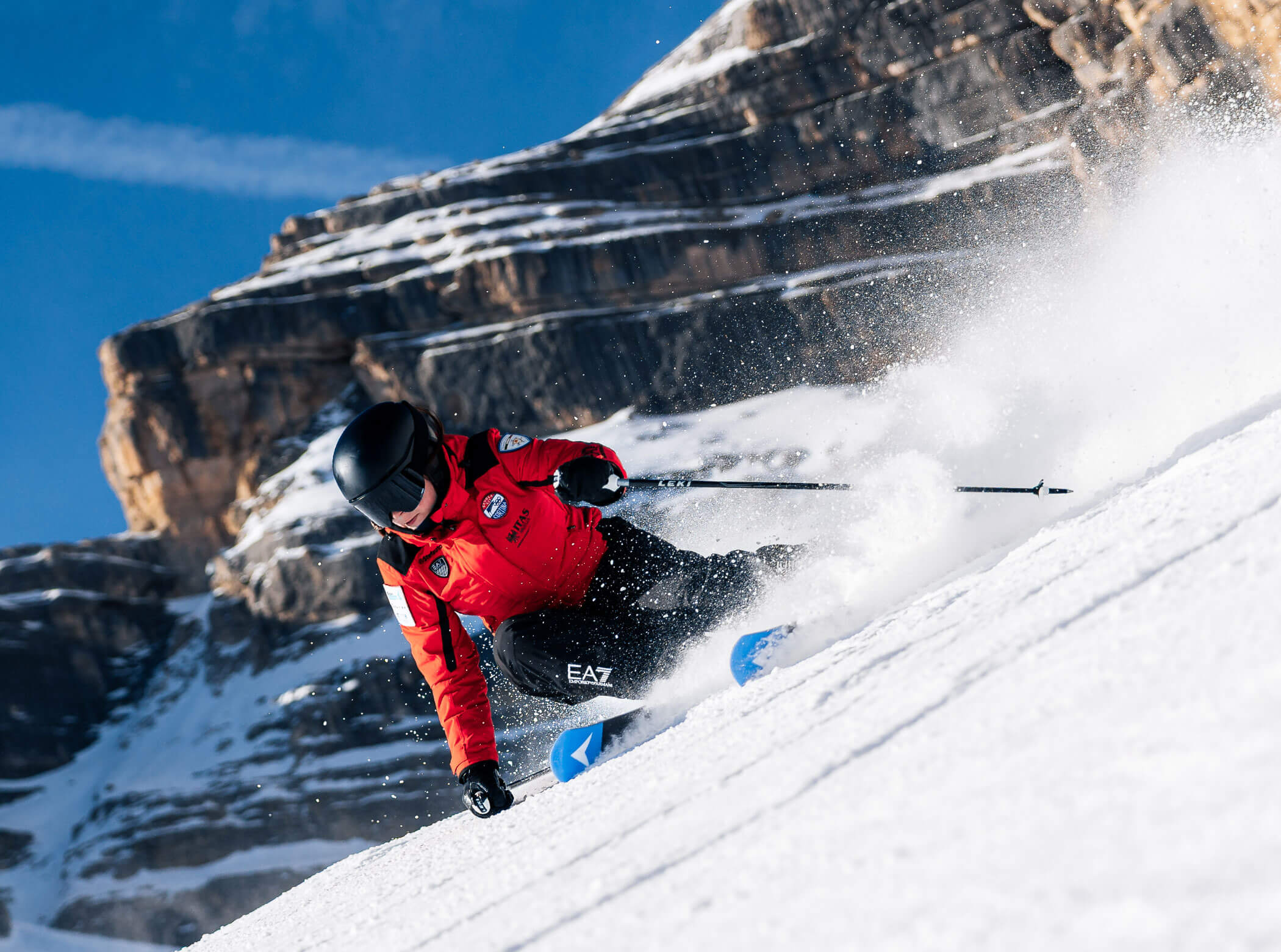 Skier in a red jacket carves quickly down a snowy slope with rocky mountains in the background. - Scuola Sci Cortina