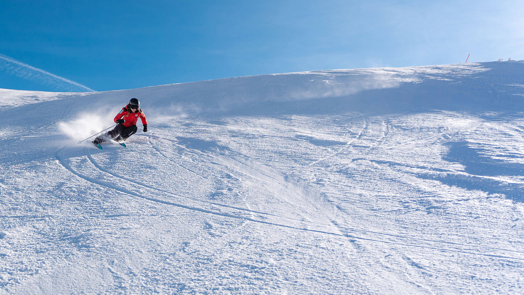 Skier in a red jacket carving down a snowy slope under a clear blue sky. - Scuola Sci Cortina
