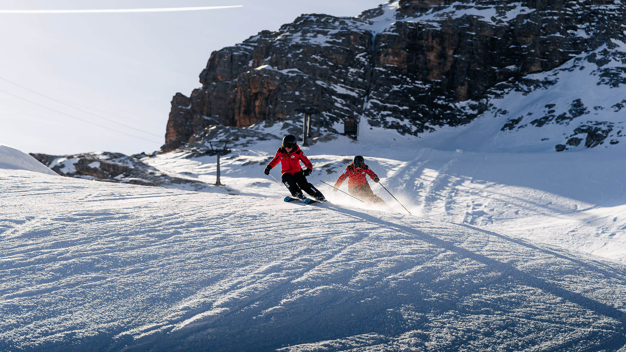 Two people in red jackets ski down a snowy mountain slope with rocky cliffs in the background. - Scuola Sci Cortina