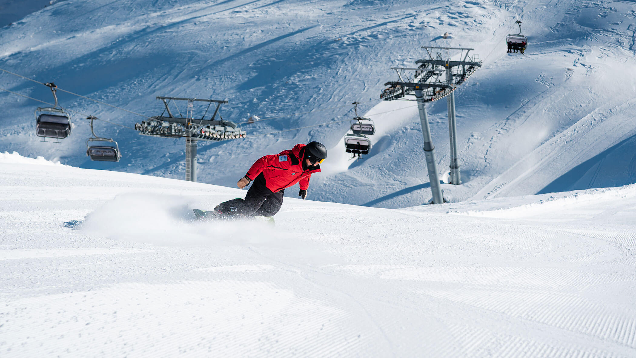 Snowboarder in a red jacket rides down a snowy slope near ski lifts on a sunny winter day. - Scuola Sci Cortina