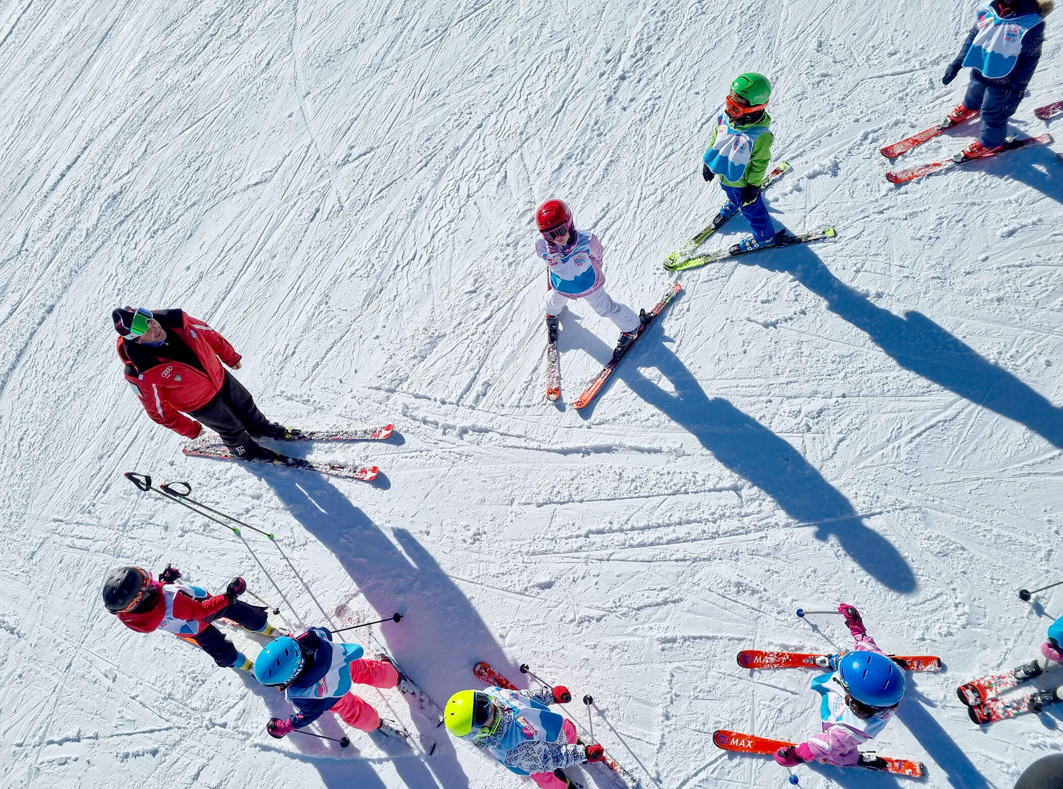 Ski instructor and kids in bright helmets stand on snowy slope, seen from above, casting long shadows. - Scuola Sci Cortina