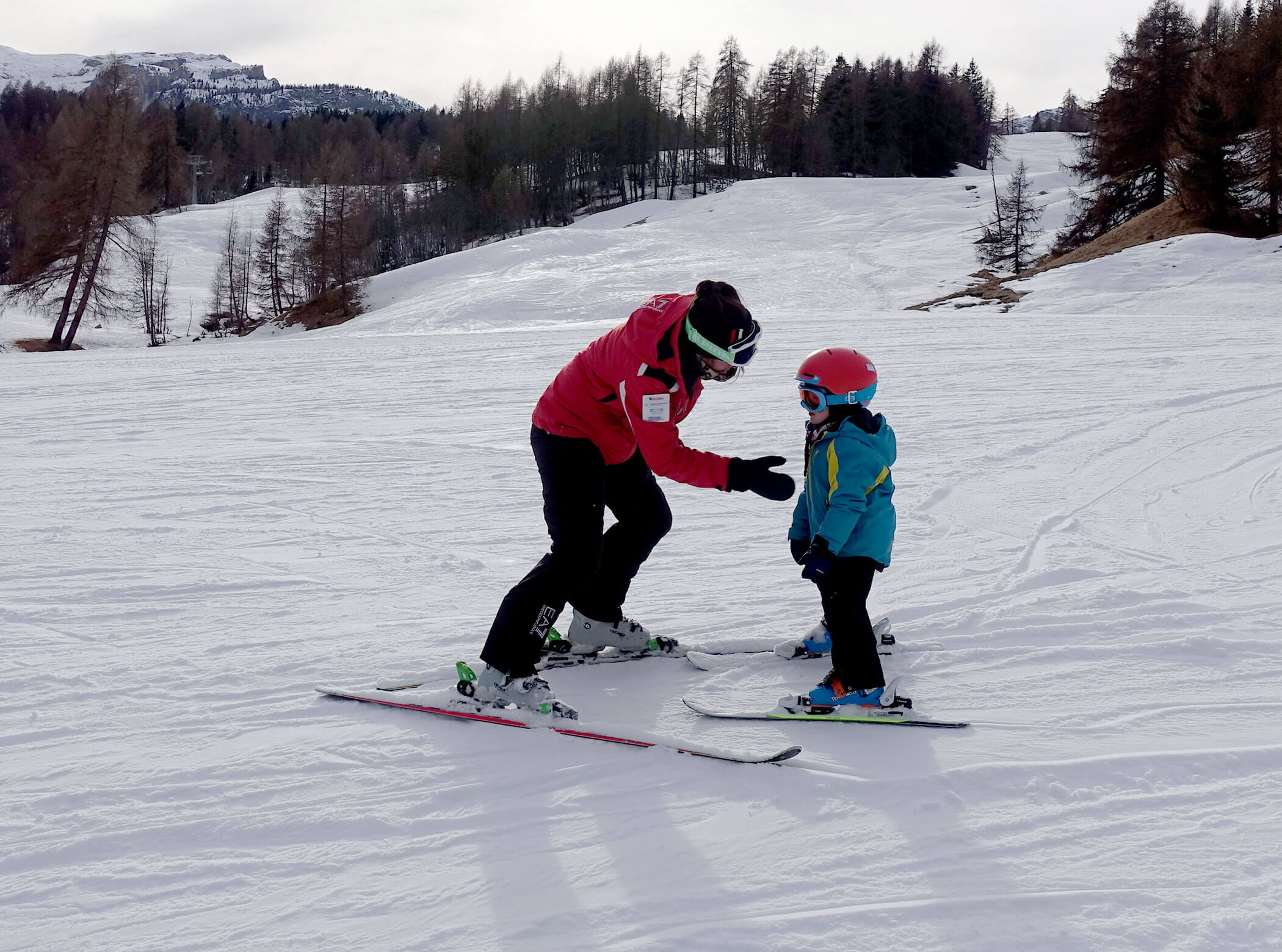 An adult teaches a child to ski on a snowy slope, both wearing helmets and winter gear. - Scuola Sci Cortina