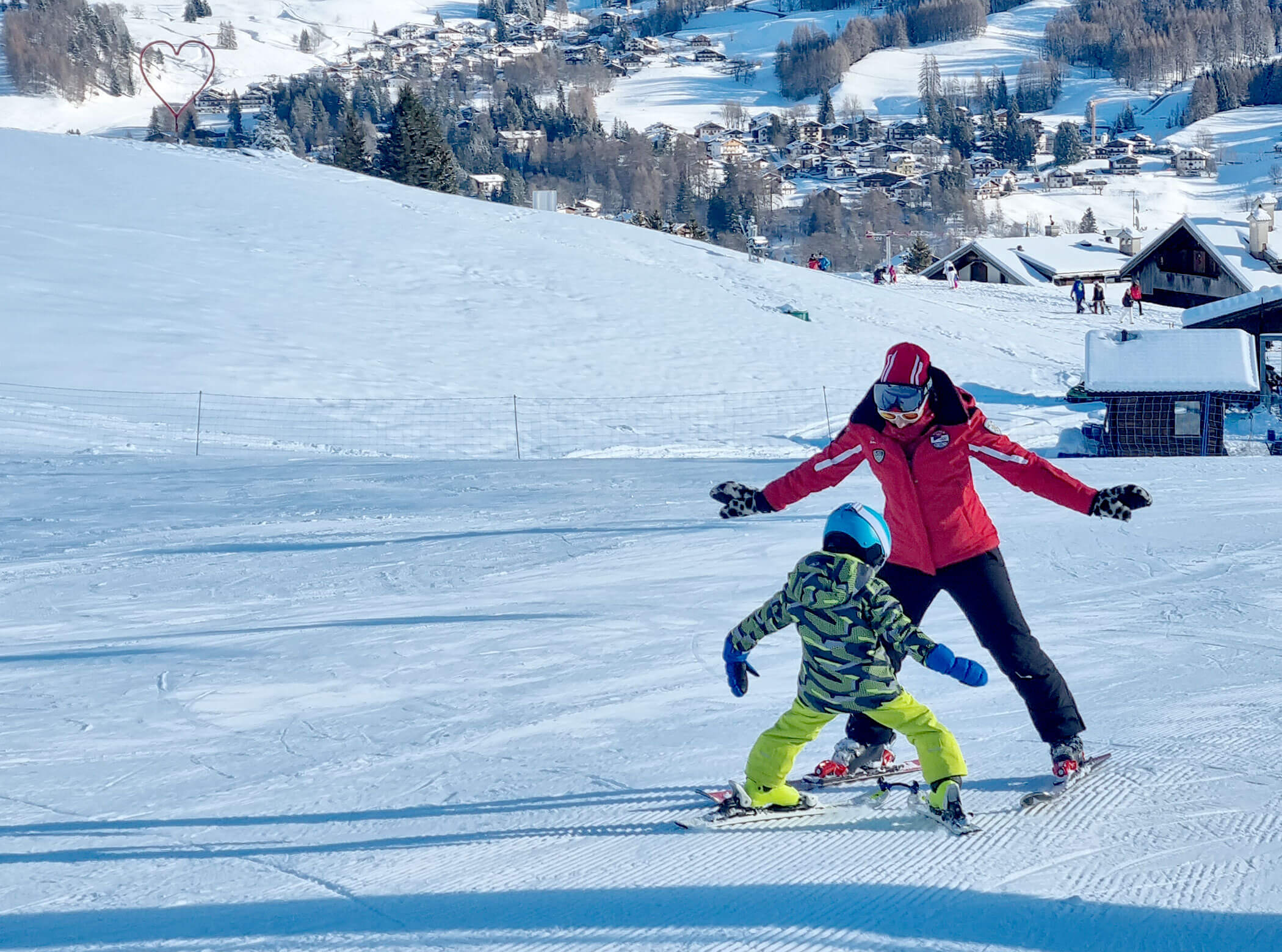 Adult teaching a child to ski on a snowy slope, with chalets and trees in the background. - Scuola Sci Cortina
