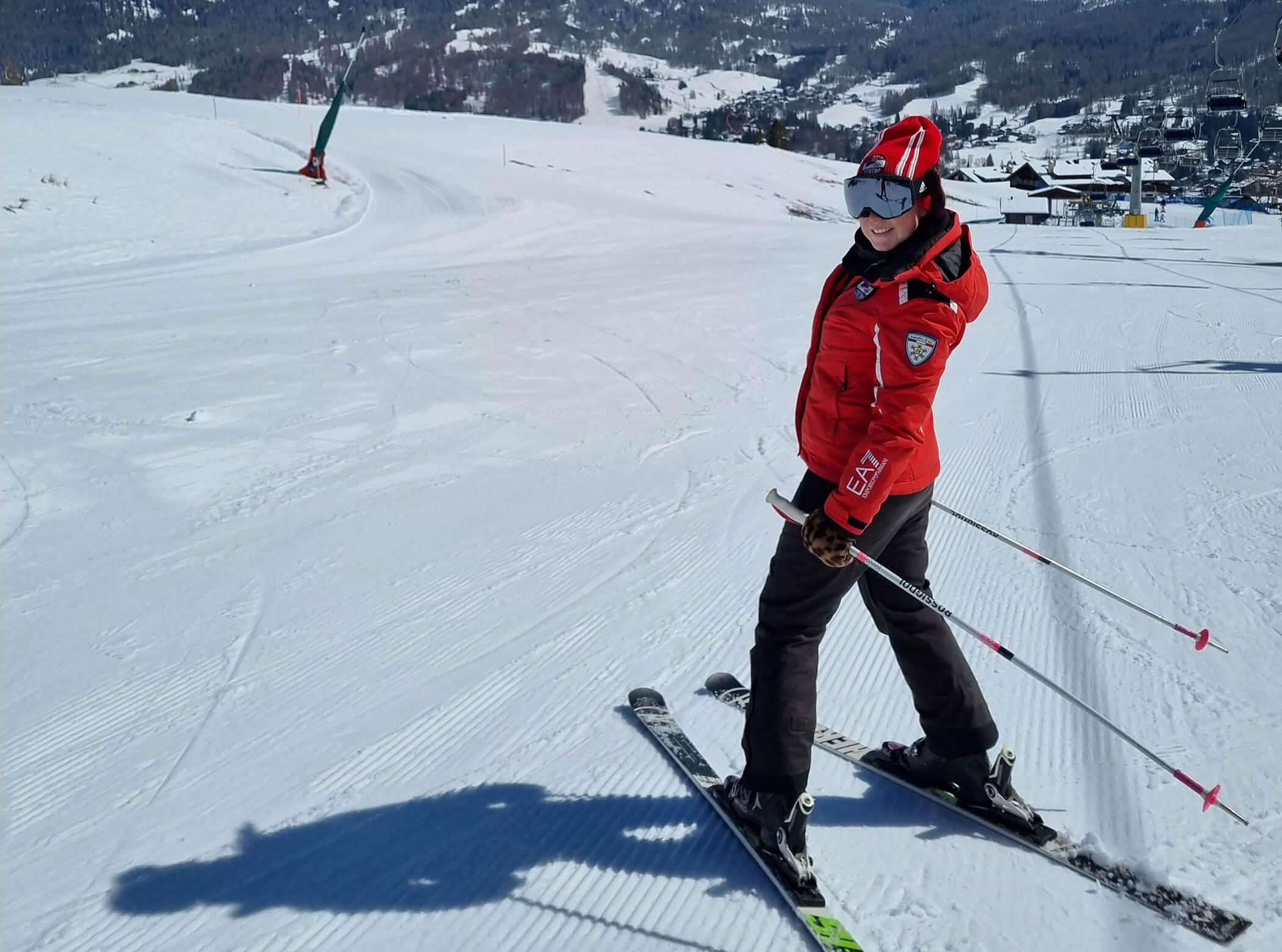 Person in red jacket skiing on a sunny, snowy slope with mountains and buildings in the background. - Scuola Sci Cortina