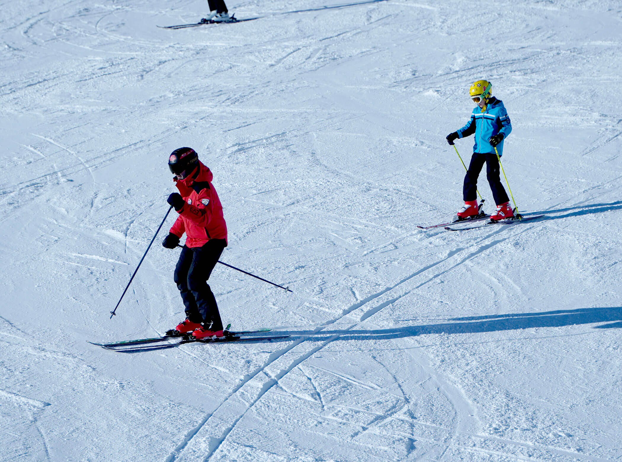 Two skiers, one in red and one in blue, ski down a snowy slope on a sunny day. - Scuola Sci Cortina