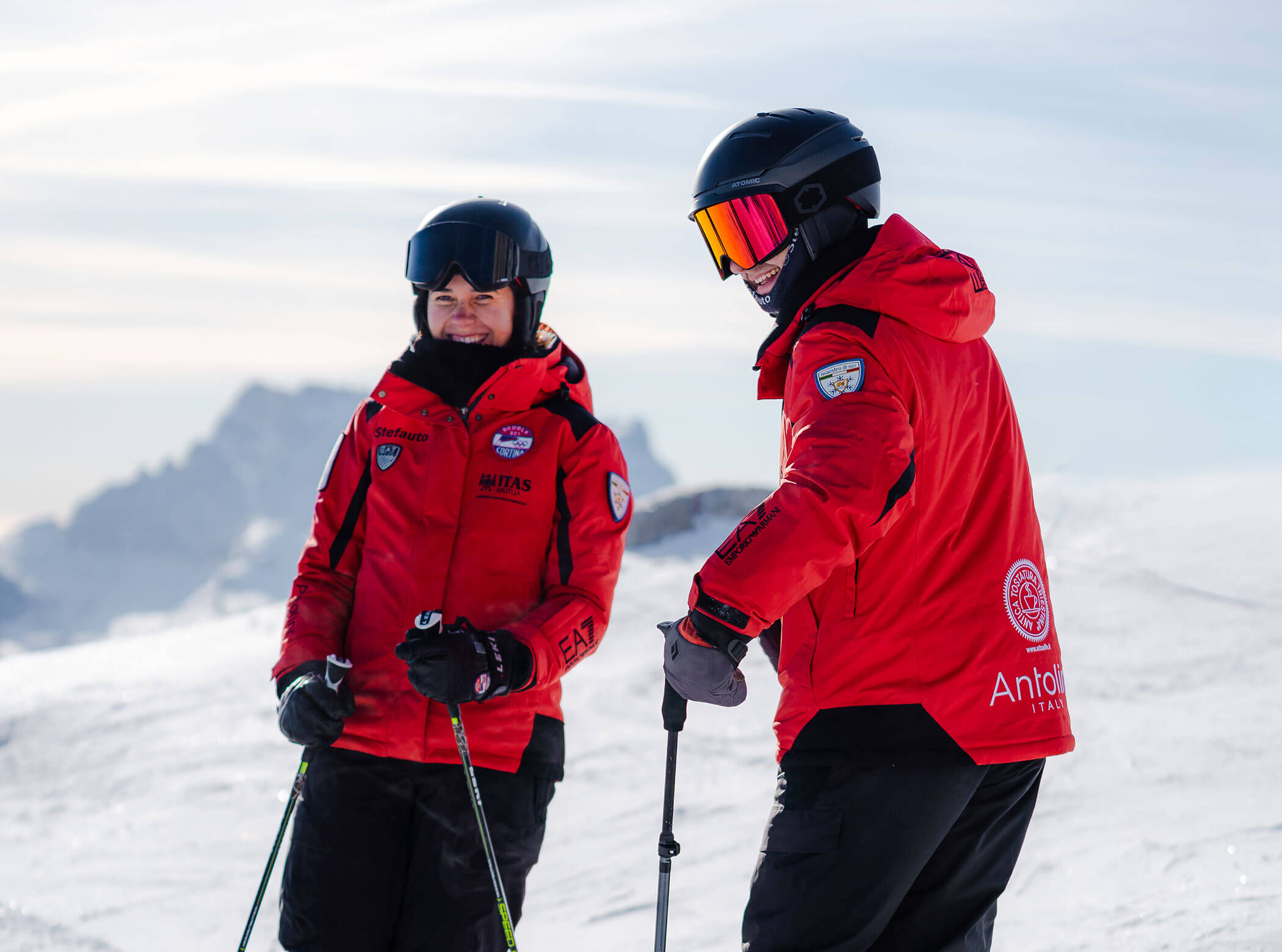 Two skiers in red jackets stand on a snowy slope, smiling and holding ski poles, mountains in background. - Scuola Sci Cortina