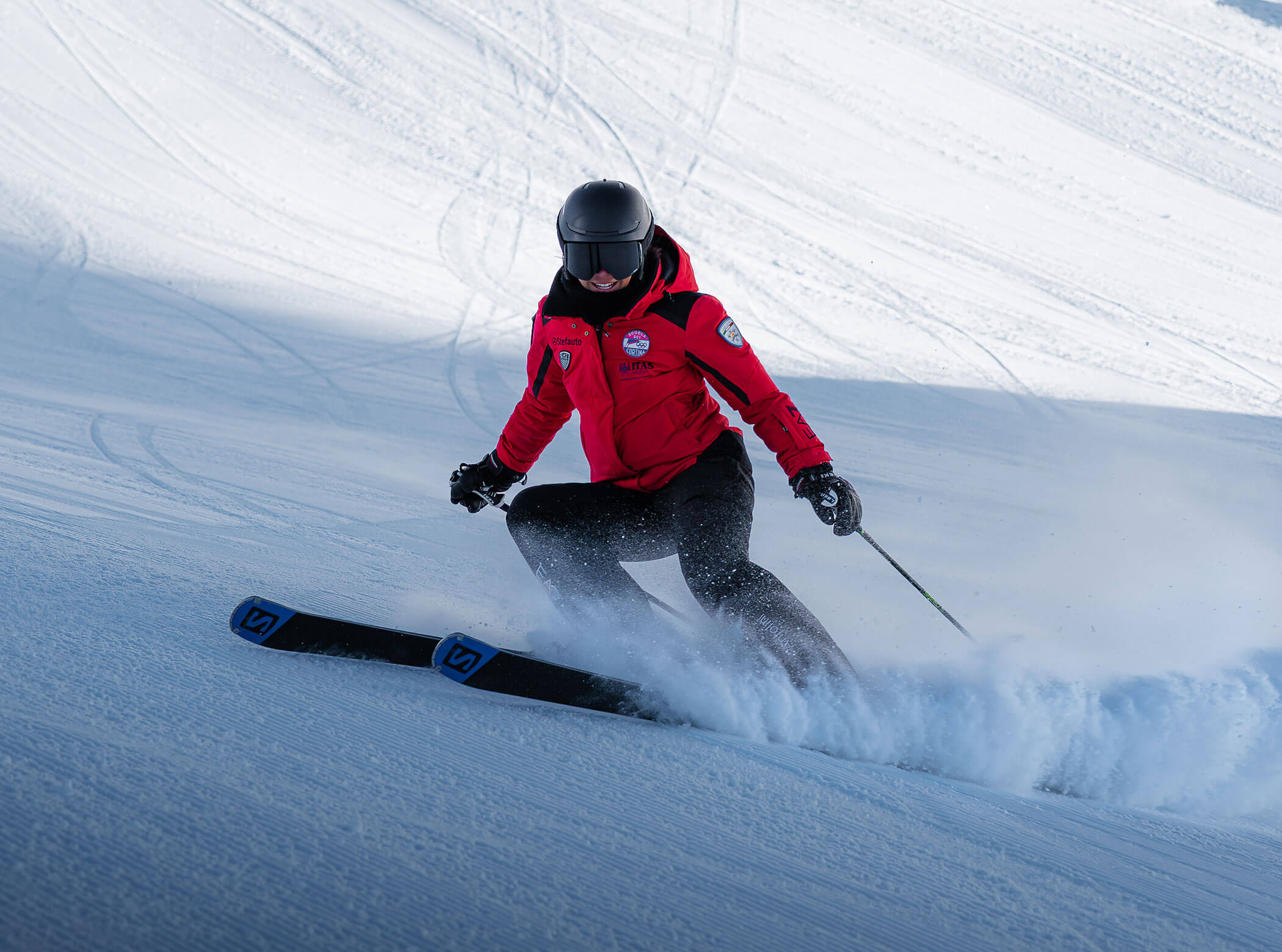 A skier in a red jacket and black helmet skis downhill on a snowy slope, kicking up powder. - Scuola Sci Cortina