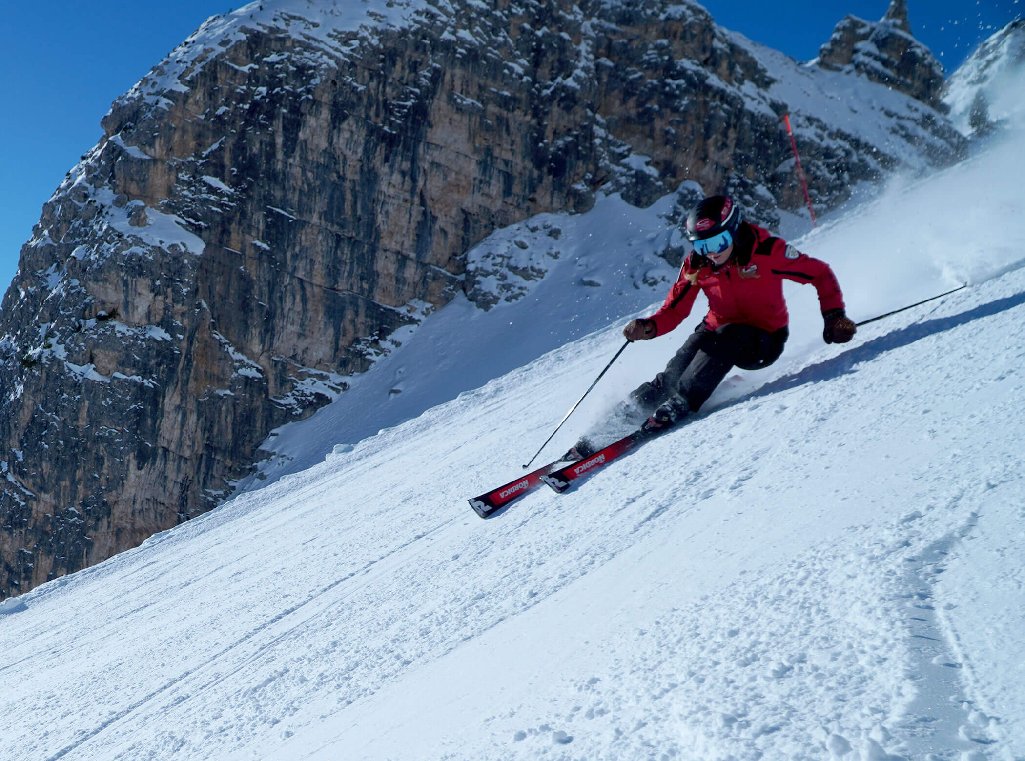 A skier in a red jacket speeds down a snowy mountain slope with rocky cliffs in the background. - Scuola Sci Cortina