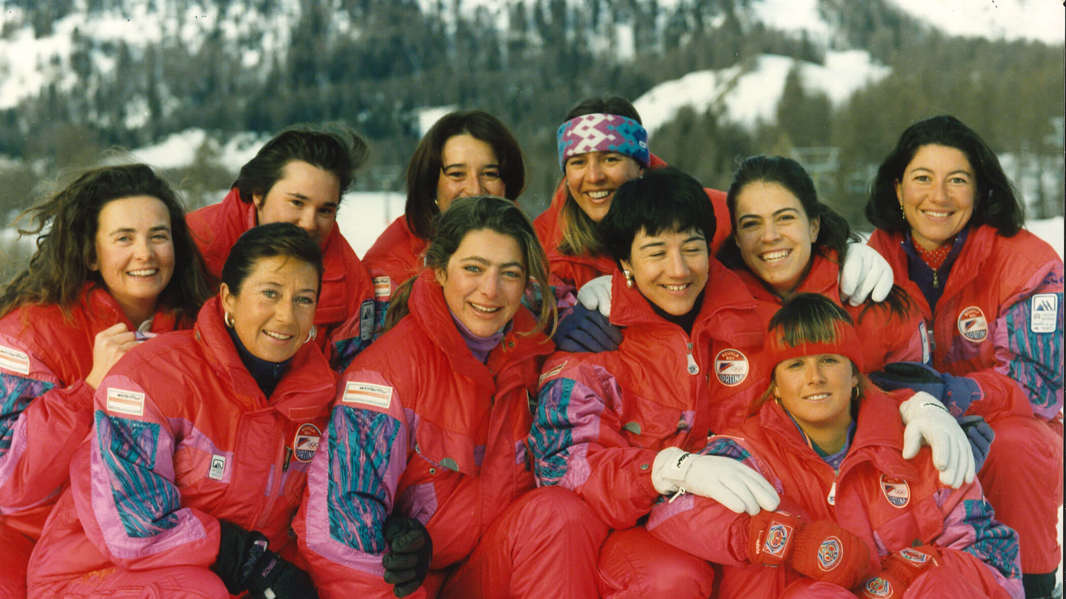 Ten women in matching red and purple ski suits pose together, smiling, with snowy mountains in the background. - Scuola Sci Cortina