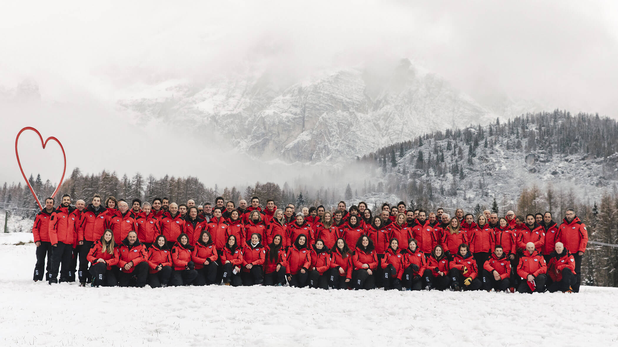 Large group in red jackets pose on snowy ground with mountains, trees, and a red heart outline in the background. - Scuola Sci Cortina