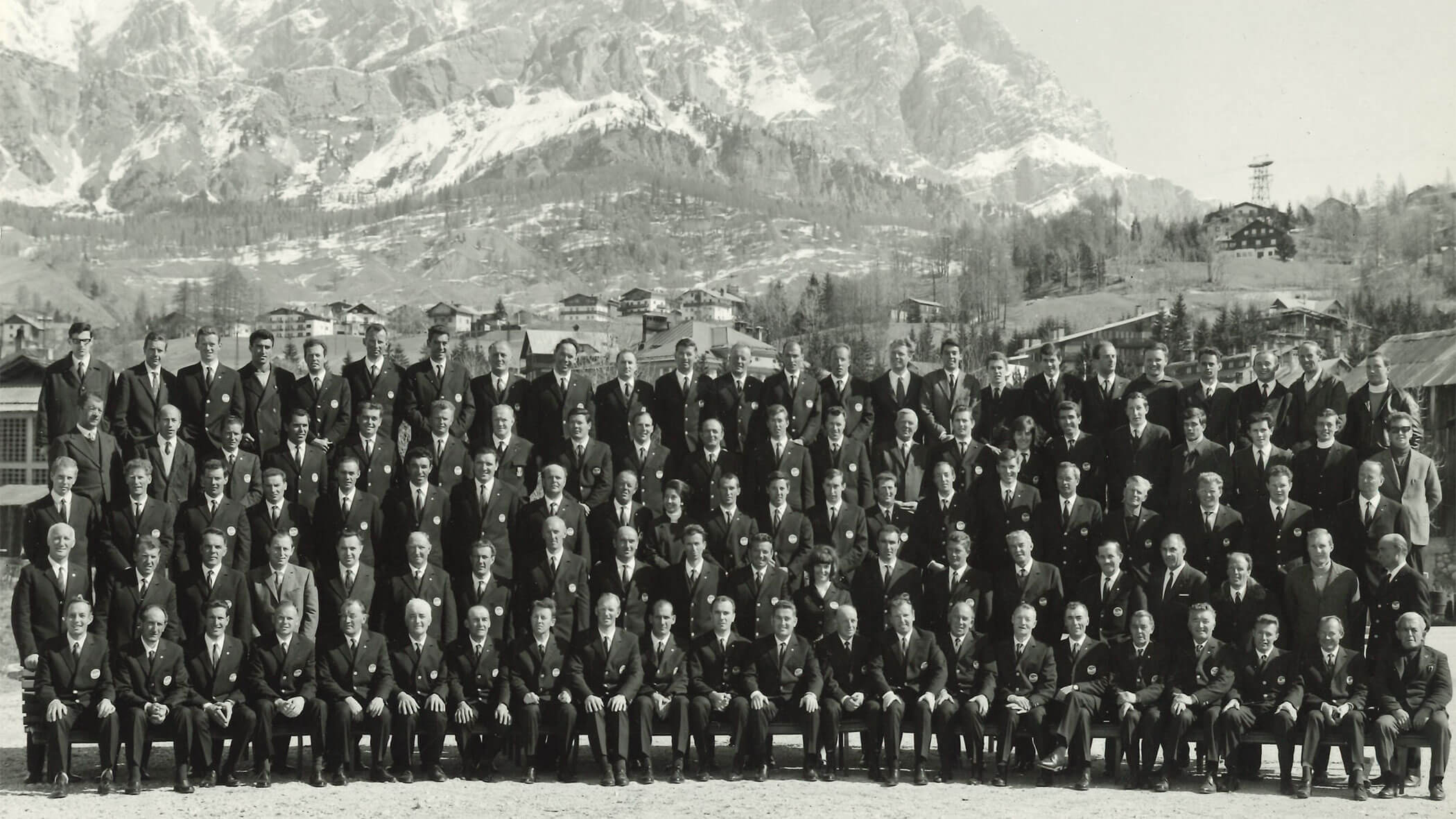 Large group of men in suits posed outdoors with snowy mountains and buildings in the background. - Scuola Sci Cortina