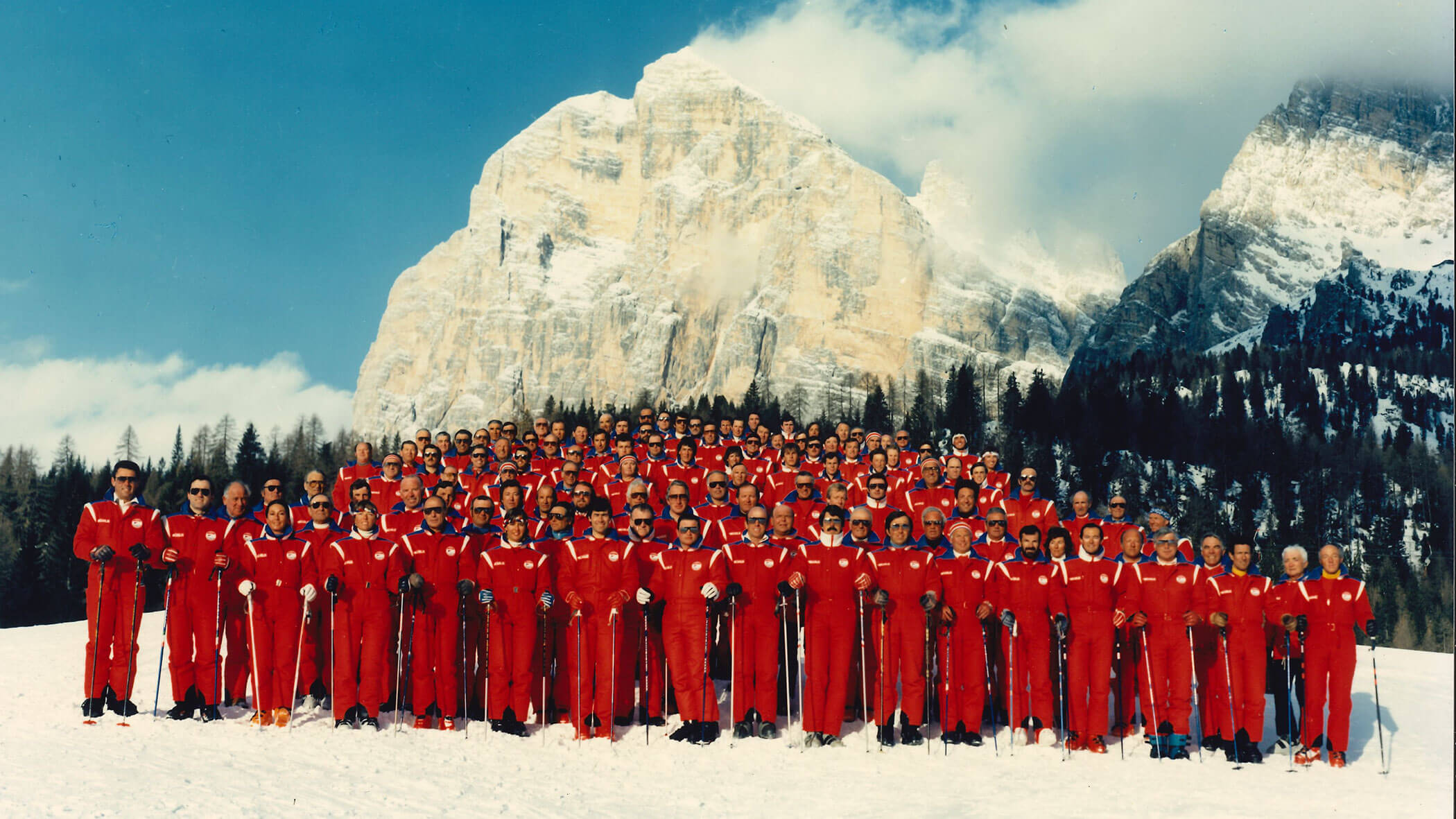 A large group in matching red ski suits poses on snowy ground with mountains in the background. - Scuola Sci Cortina